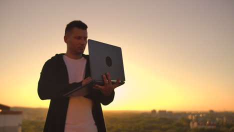 Ein-Mann-Mit-Einem-Laptop-Steht-Bei-Sonnenuntergang-Auf-Dem-Dach-Des-Gebäudes-Und-Schreibt-Einen-Code-Auf-Der-Tastatur.-Hacker-Mit-Laptop.